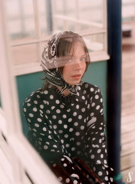 Young woman in a black-and-white polka dot shirt and paisley headscarf poses indoors near a window with diffused natural light.