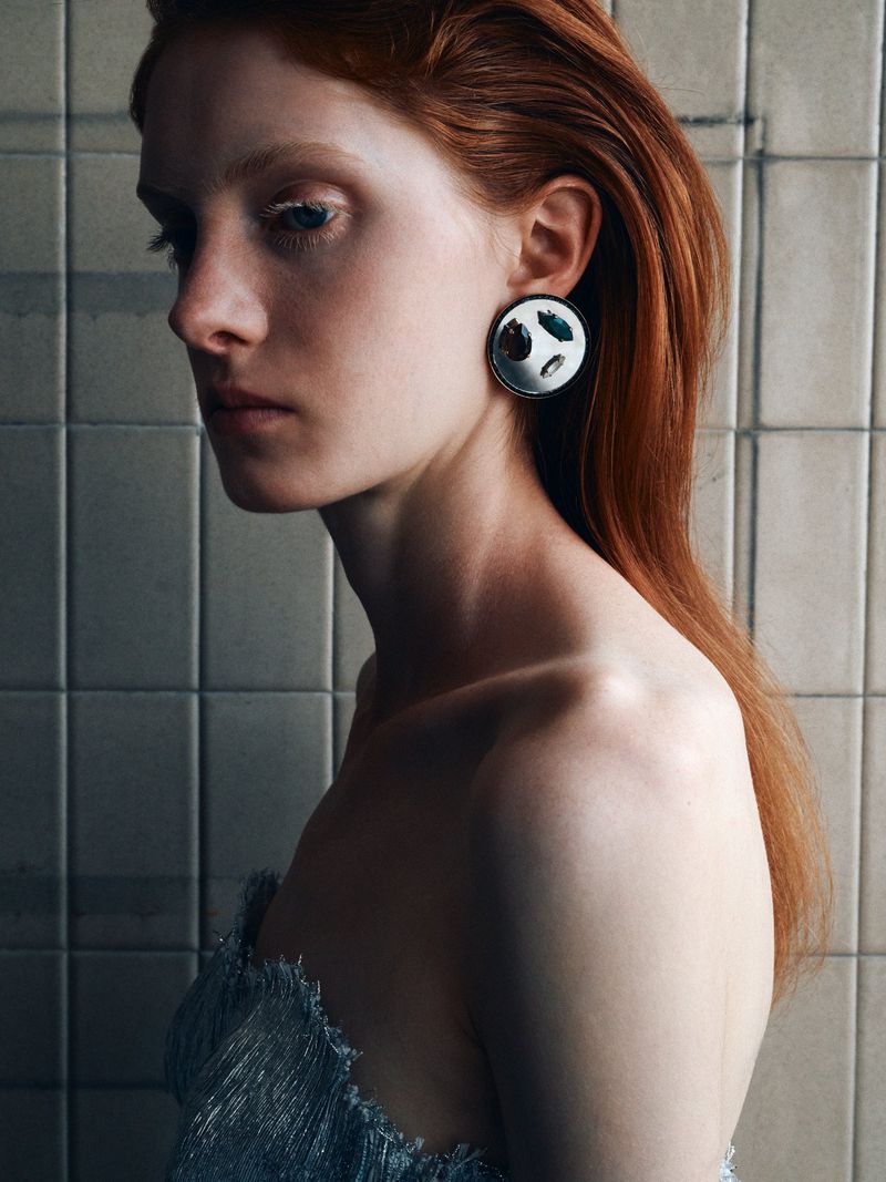 Close-up portrait of a young red-haired woman with fair skin wearing a silver and white earring in front of a tile wall.