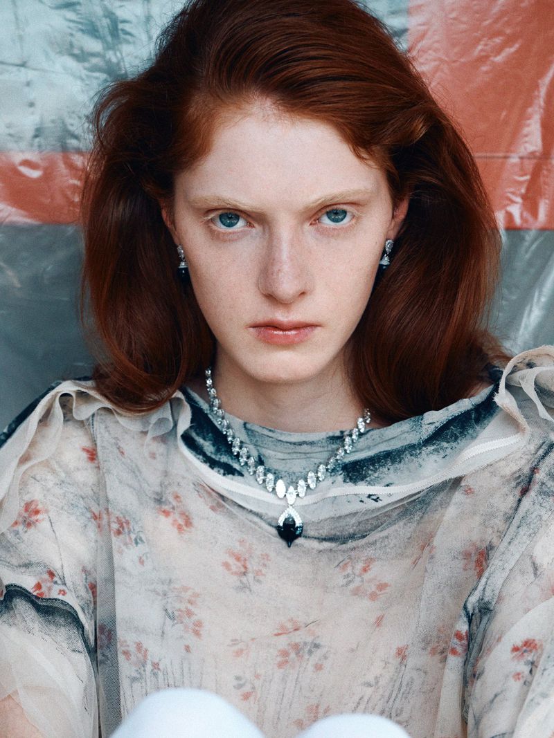 Close-up studio portrait of a red-haired woman with pale skin wearing vintage floral top and gemstone jewelry looking directly at the camera.