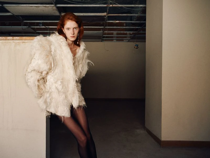 Red-haired fashion model in an oversized white fur coat leans against a wall in a dimly lit, unfinished interior space with a cement floor.