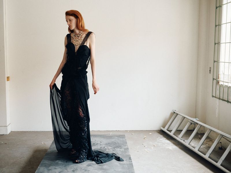A fashionable, red-haired woman wearing a long black dress and decorative necklace stands on a gray platform in an empty room with a ladder.