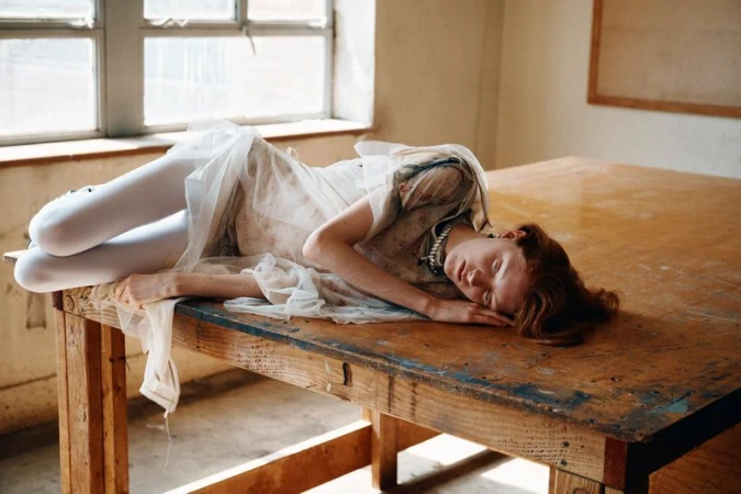 Young woman with red hair in white dress and tights reclines on an old wooden table in a sunlit room, eyes closed, daydreaming.