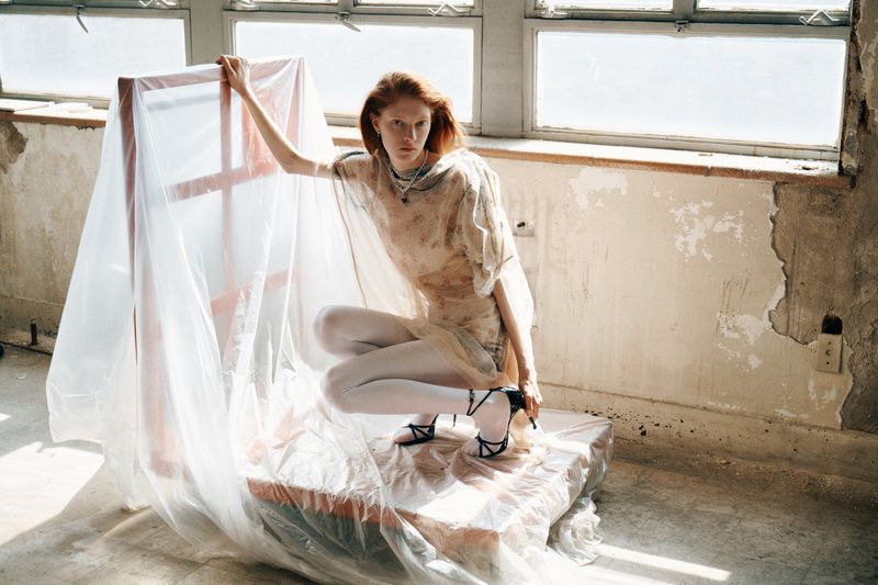 Redhead model in sheer top and white tights poses on a box covered in plastic wrap indoors with bright window lighting.