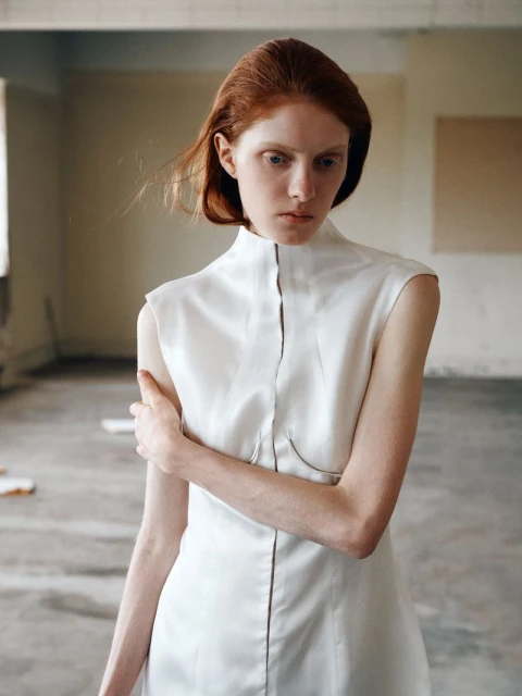 A red-haired woman in a white dress stands in an empty room with concrete floors and white walls, bathed in soft light.