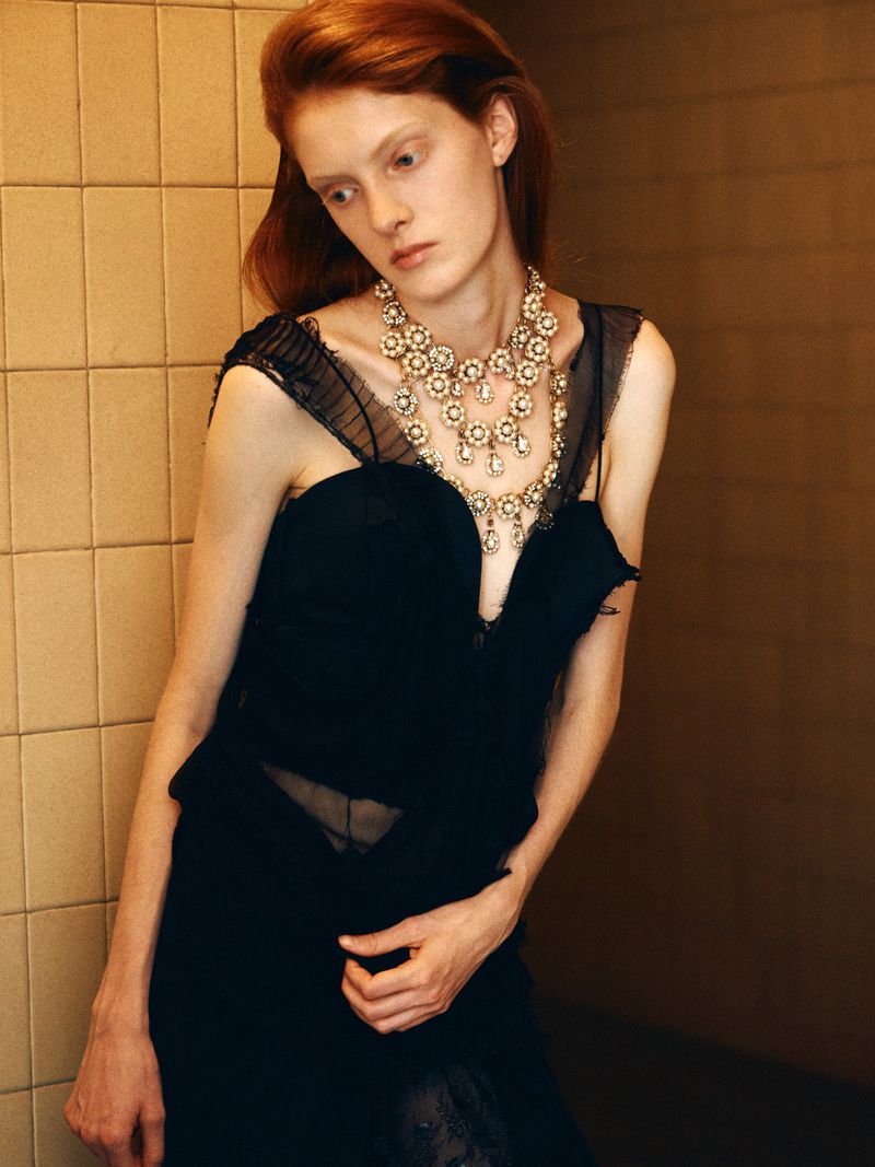 Redhead woman poses in a black dress and elaborate necklace, standing against a tiled wall in a high-fashion editorial style.