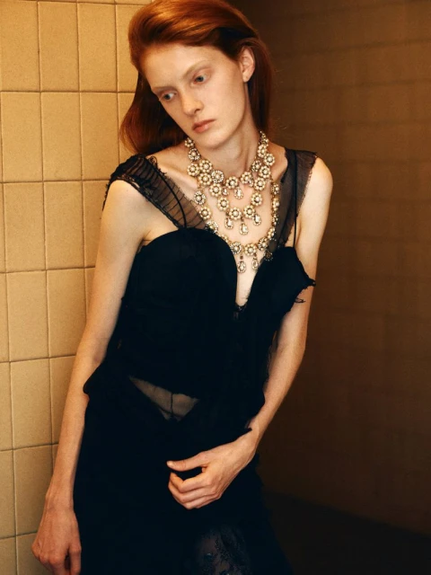 Redhead woman poses in a black dress and elaborate necklace, standing against a tiled wall in a high-fashion editorial style.