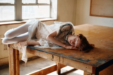 A woman in a sheer white dress lying down on a large wooden work table in an empty room with soft natural daylight.