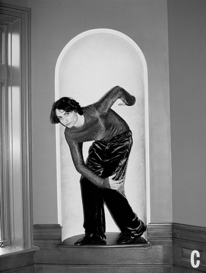 A young adult with dark hair poses in a fitted long-sleeve shirt and velvet pants, within an architectural alcove, set against a neutral interior space.