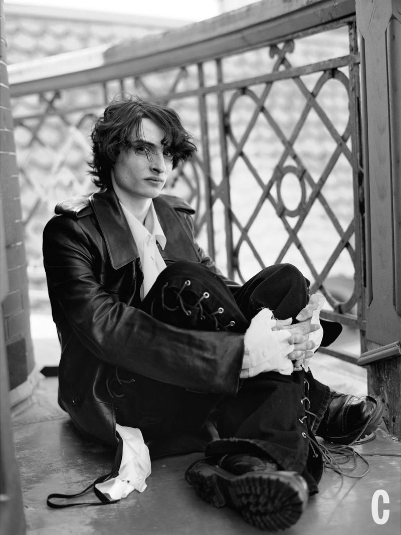 Black-and-white portrait of a young adult sitting outdoors wearing a leather jacket, lace-up pants, boots, and white gloves near an iron railing.