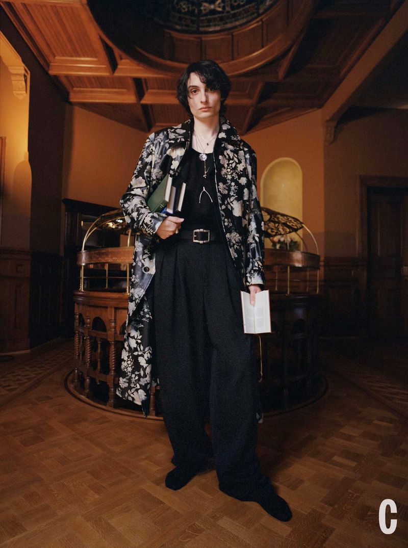 Young adult in floral coat holding books in a vintage interior setting with wood floors, ornate desk, and detailed ceiling.