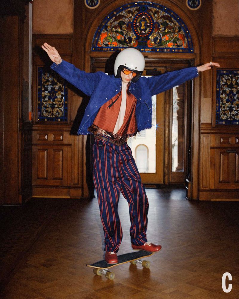 Adult male wearing a helmet, jacket, striped trousers, and shoes while riding a skateboard in an ornate indoor setting.