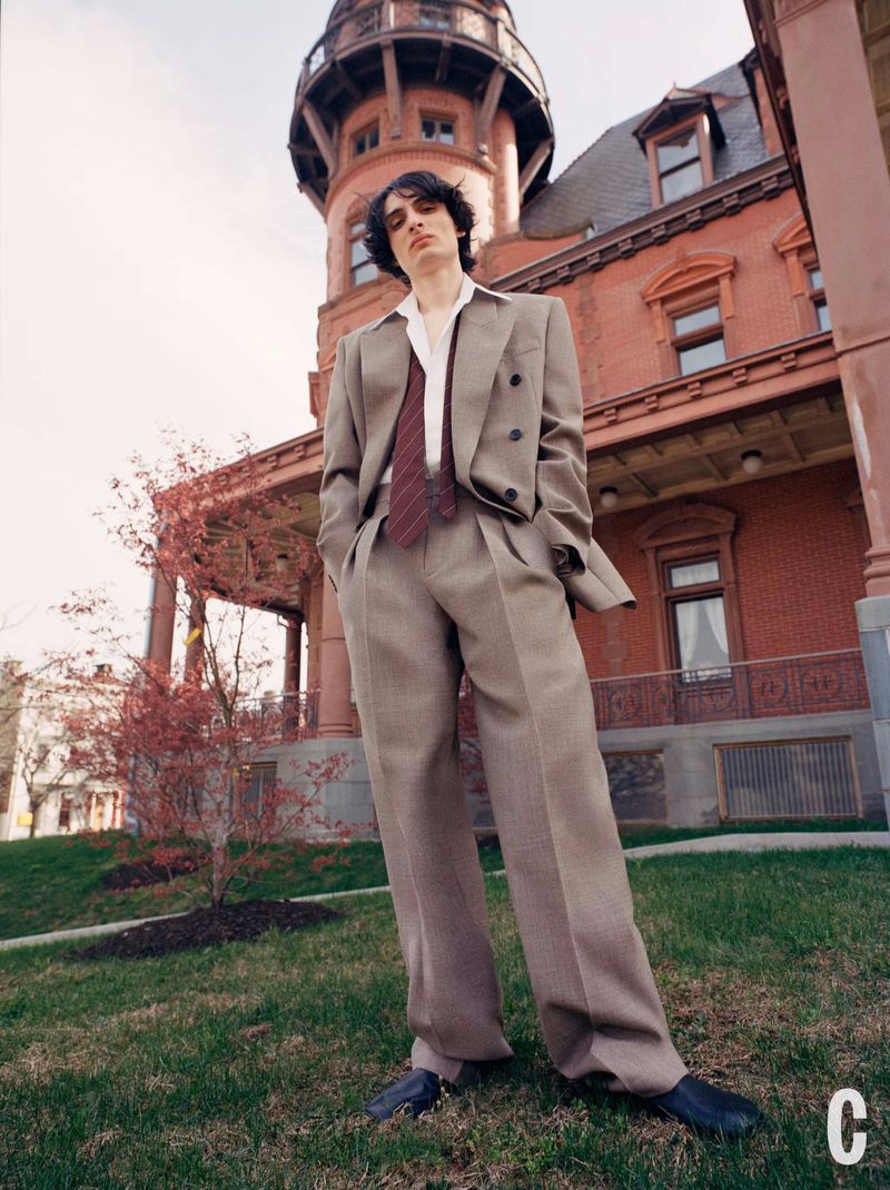Young adult poses in a tan suit against a red brick building with a turret, set on a grassy lawn beneath a bright sky.
