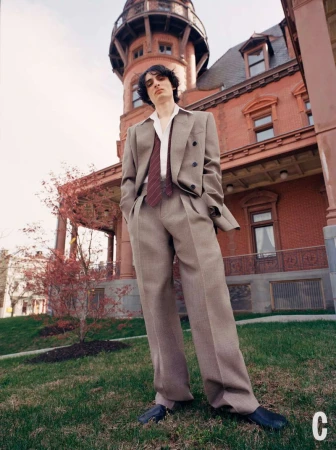 Young adult poses in a tan suit against a red brick building with a turret, set on a grassy lawn beneath a bright sky.