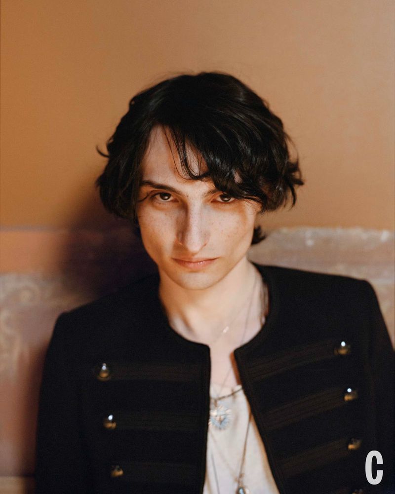 Portrait of a young adult with dark hair, wearing a black jacket and necklaces, set against a muted brown background with soft lighting.