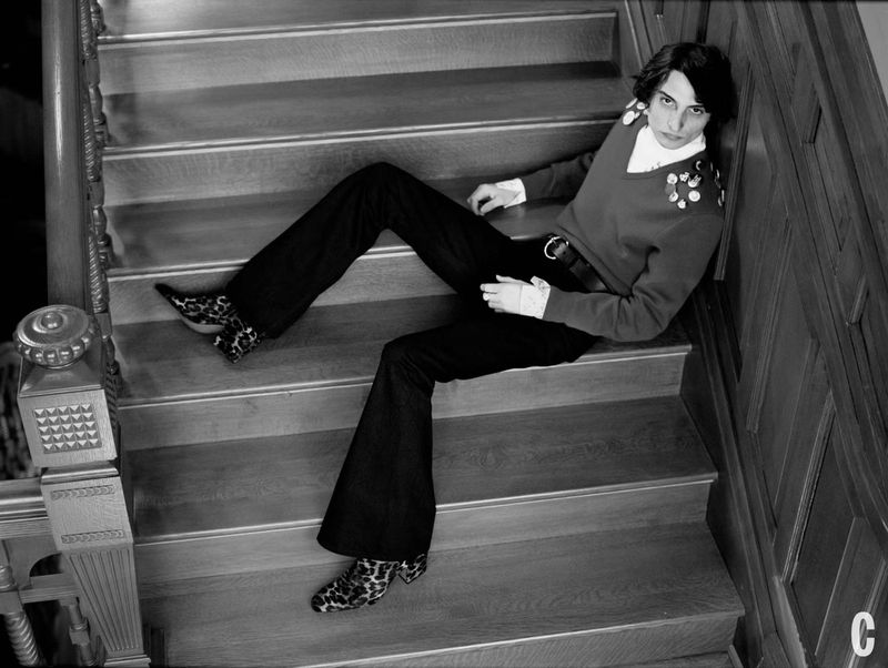 Black and white portrait of a person with dark hair and a red sweater lying on a wooden staircase with patterned heeled shoes.