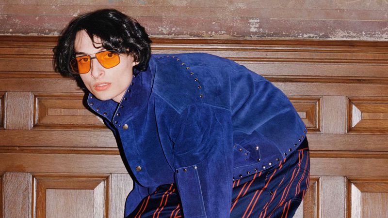 Teenager in an embellished blue jacket and striped pants poses indoors in front of an ornate paneled wall, wearing orange-tinted sunglasses.