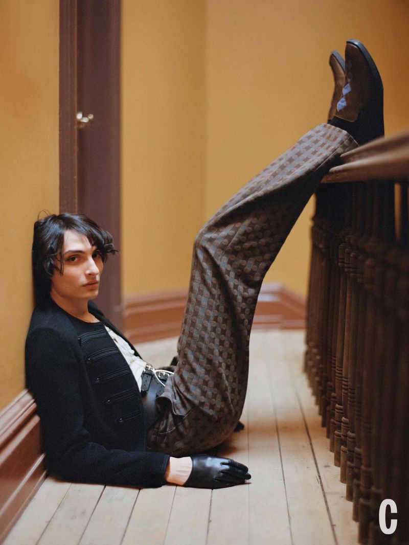 Young adult poses indoors with patterned pants and black jacket, legs extended to the railing. Warm, inviting color palette.