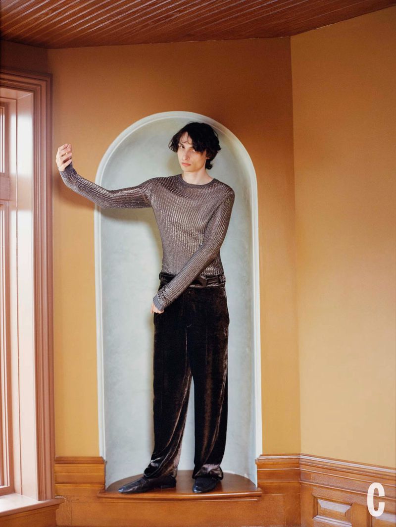A young adult poses in a niche, wearing a shimmering long-sleeved shirt and velvet pants, set against peach-toned walls with natural window light.