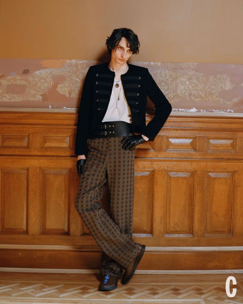 Fashionable person poses against a wooden paneled wall, wearing a dark jacket, white shirt, brown patterned pants, and leather gloves.