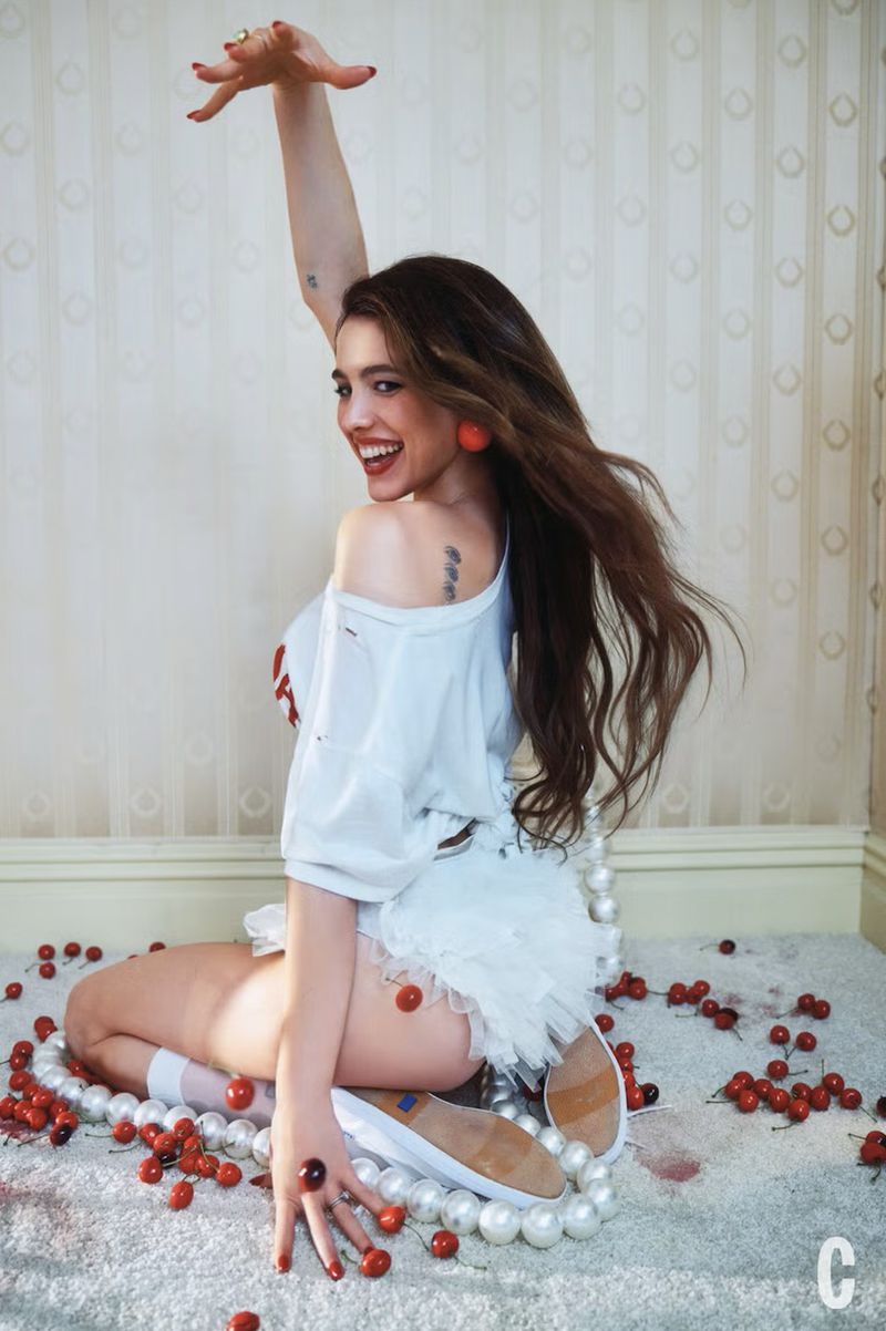 Smiling woman in a white shirt and tutu sits on a carpet with cherries and pearls, turning her head back in an indoor setting.