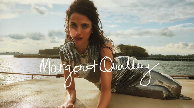 Margaret Qualley poses in a shiny silver dress against a cloudy coastal backdrop. Fashion shoot, outdoor setting, model looking at camera.