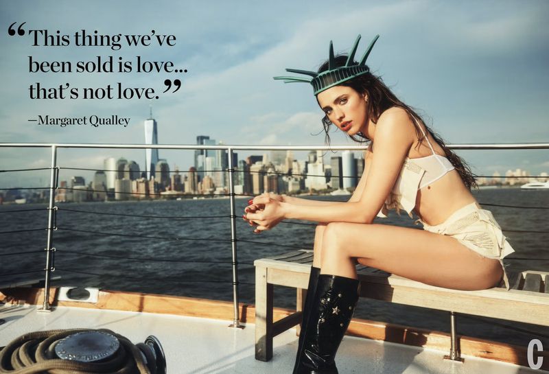 Fashion shot on a boat deck: a woman wearing a Statue of Liberty crown, posing against a city skyline and ocean backdrop, boat details.
