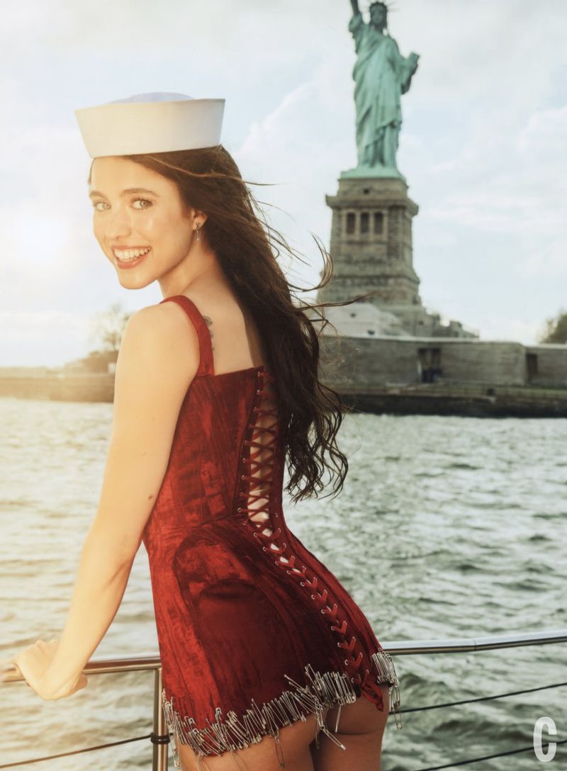 Smiling woman in a red dress and sailor hat looks over her shoulder with the Statue of Liberty in the background and shimmering water.