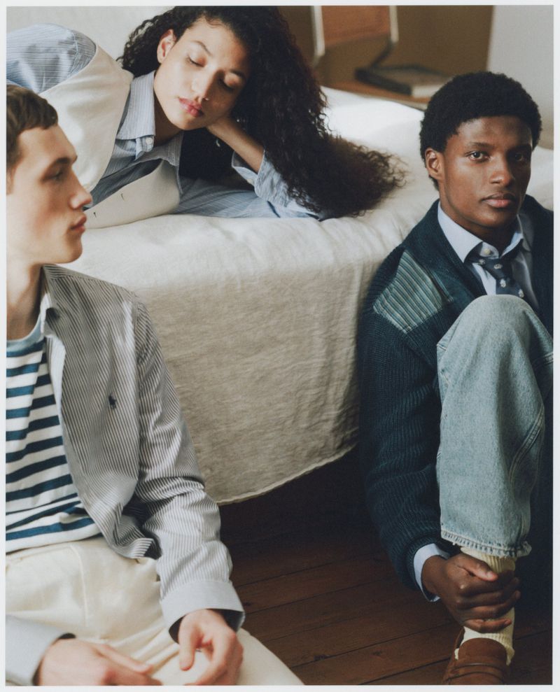 Three people seated in a room: two men in pinstripe jackets and a woman in blue shirt leaning on a bed. Natural light, casual wear.