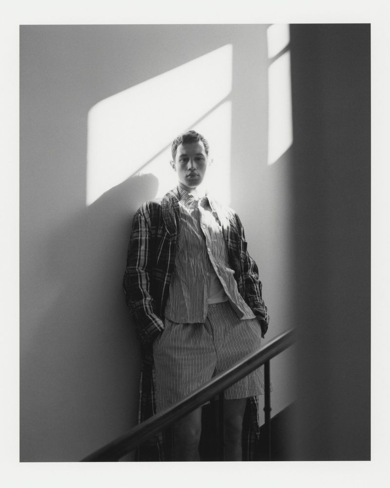 A young adult male stands on a staircase wearing shorts, a striped shirt, and a plaid jacket, with natural light casting shadows.