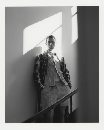 A young adult male stands on a staircase wearing shorts, a striped shirt, and a plaid jacket, with natural light casting shadows.