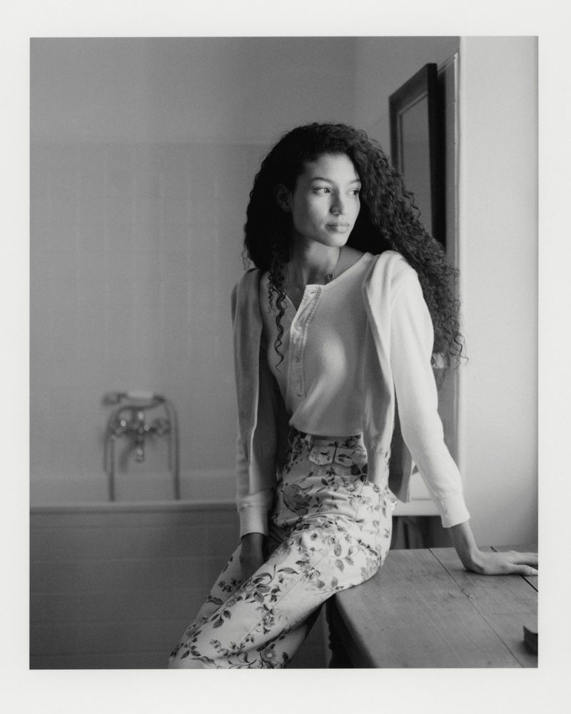 A young woman with curly hair, wearing floral print pants and a cropped top and cardigan, sits on a wooden table in a bright room.
