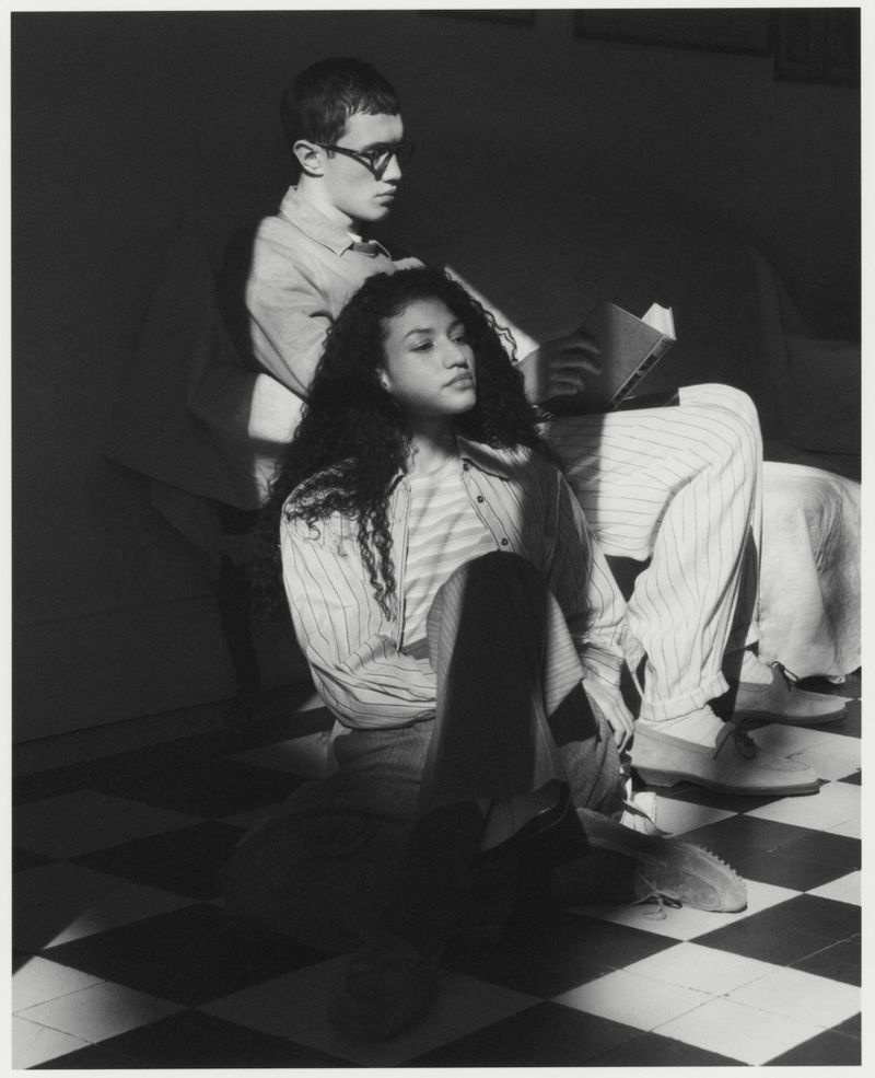 Monochrome image captures a young man reading behind a young woman with curly hair indoors on a black and white checkered floor.