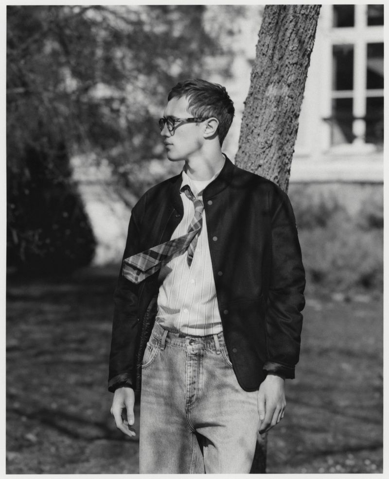 Monochrome portrait of a young adult man in a jacket, striped shirt, and jeans, leaning against a tree outside a building.