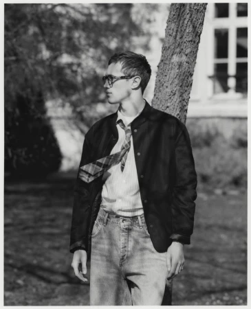 Monochrome portrait of a young adult man in a jacket, striped shirt, and jeans, leaning against a tree outside a building.
