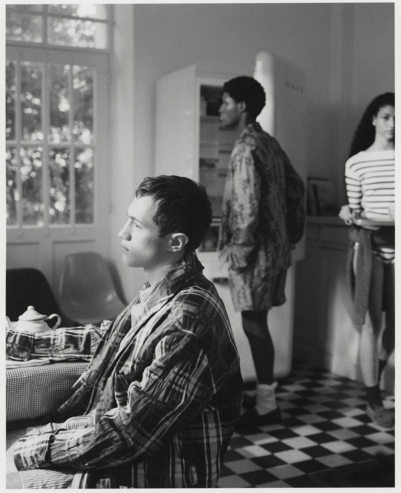 Black-and-white image of three people in a kitchen: a man in a plaid robe seated, a woman standing by a refrigerator, and a woman in a striped shirt.