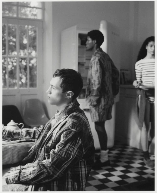 Black-and-white image of three people in a kitchen: a man in a plaid robe seated, a woman standing by a refrigerator, and a woman in a striped shirt.