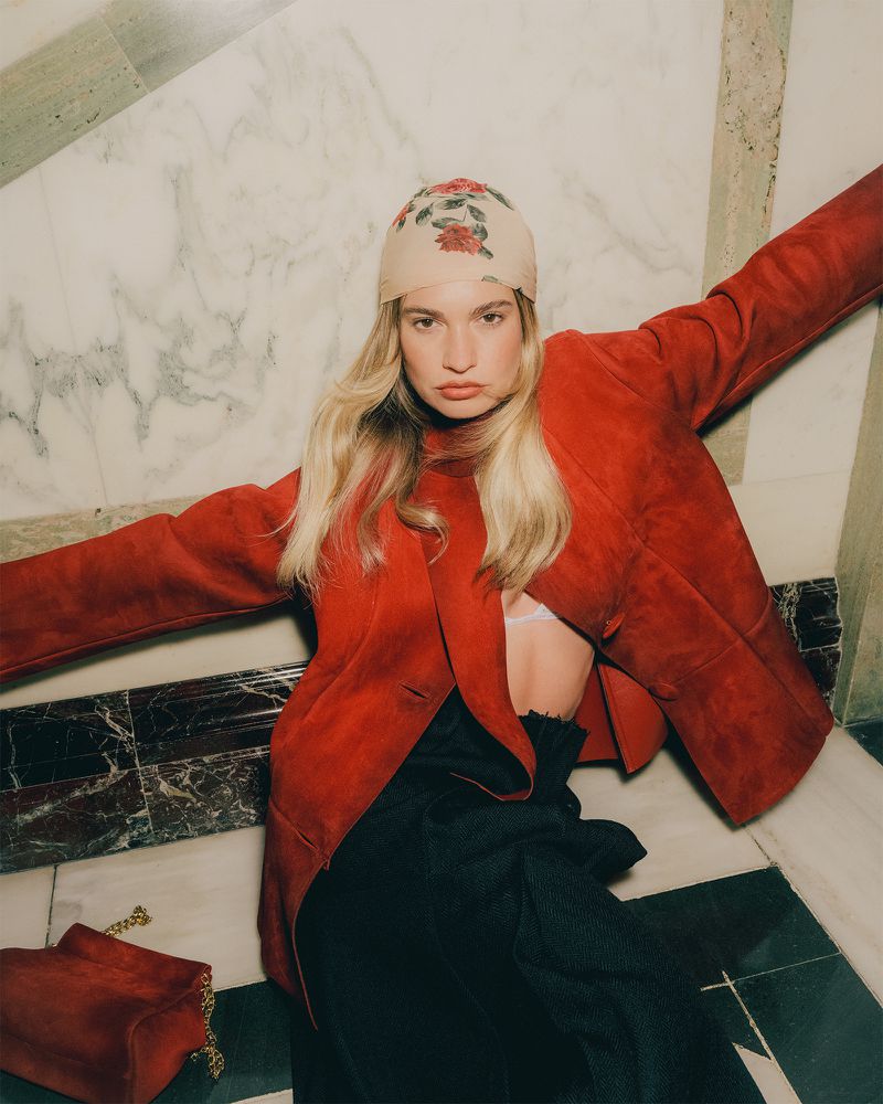 Stylish adult female poses while wearing a red jacket, headscarf, and black pants with a red purse on a marble floor.