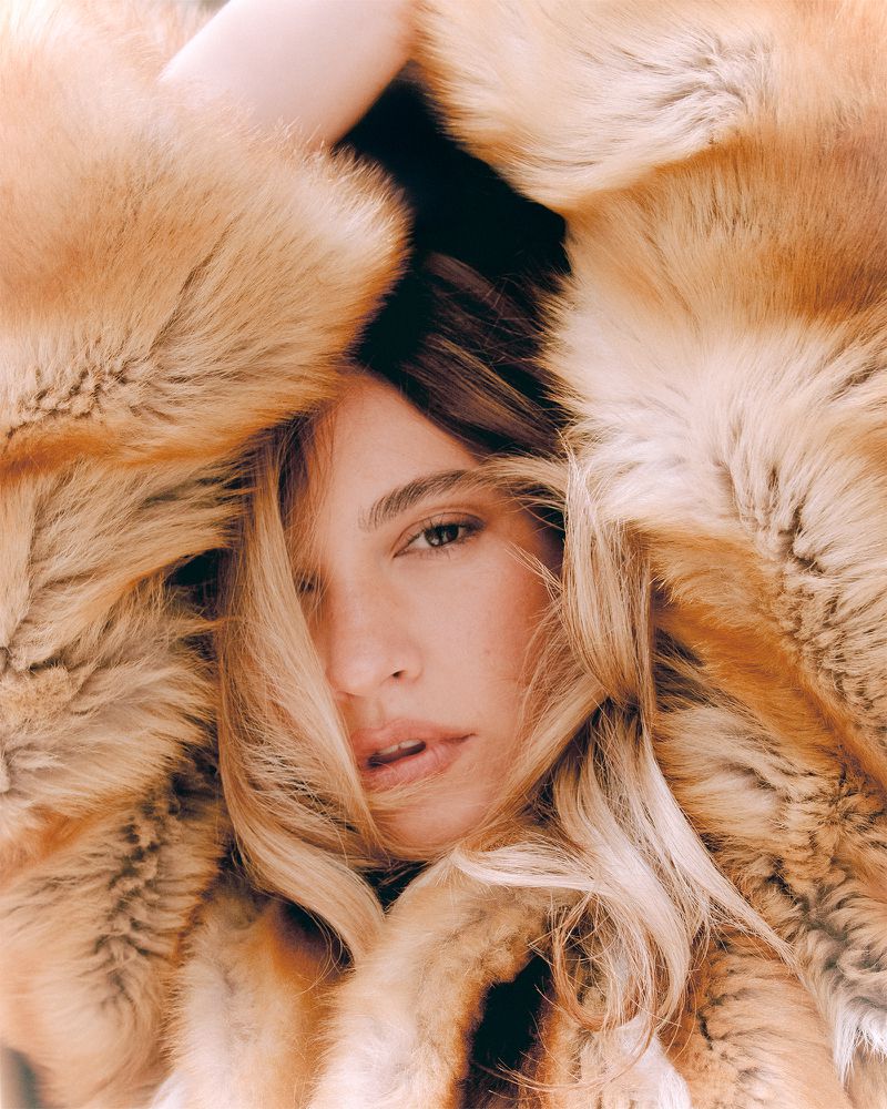 Close-up studio portrait of a woman with blonde hair and light skin, posing with a fluffy brown fur coat framing her face.