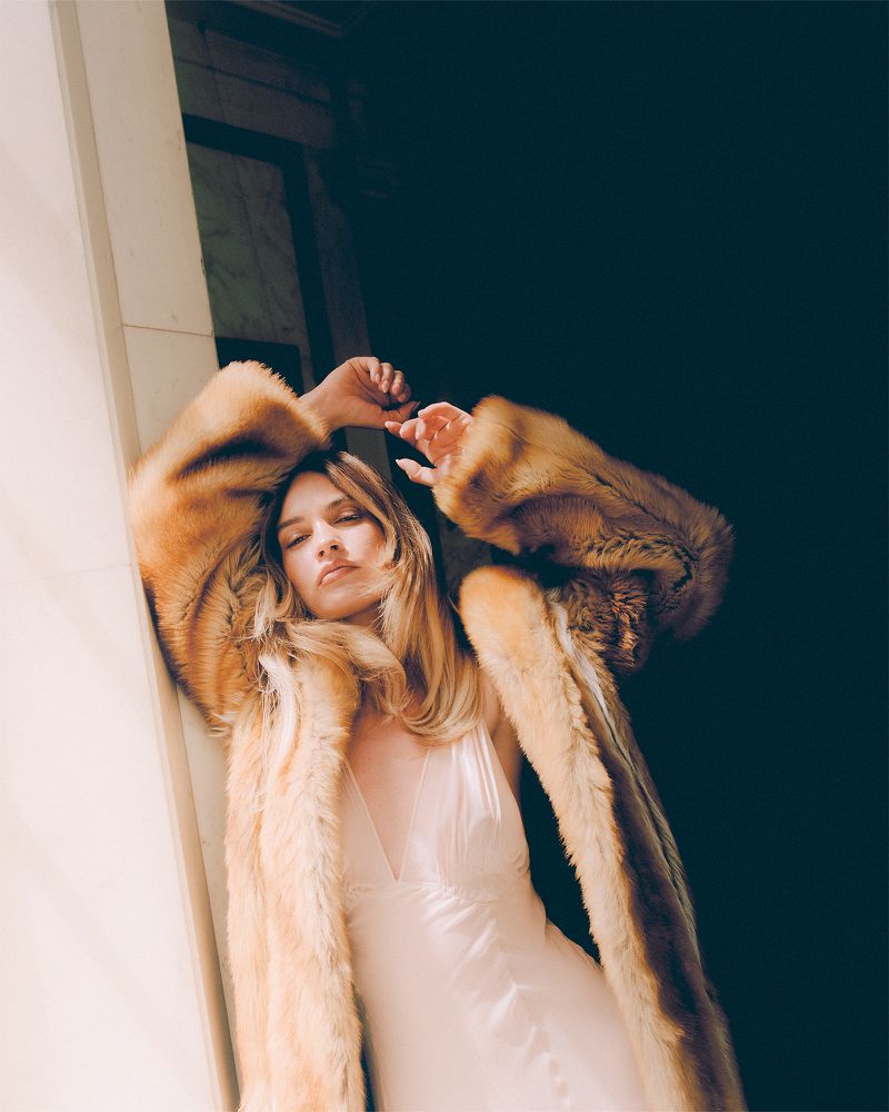 Blonde woman in a dress and brown fur coat leans against a white doorframe in a warmly lit indoor setting with dark background.