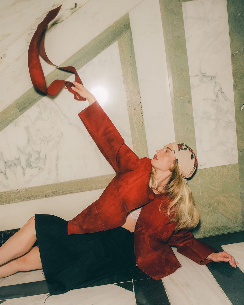 Woman in red jacket lying on marble floor, holding red ribbon. Blonde hair, patterned headscarf. Fashion, beauty, indoor shot.