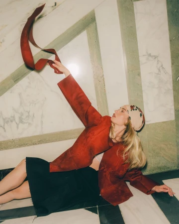 Woman in red jacket lying on marble floor, holding red ribbon. Blonde hair, patterned headscarf. Fashion, beauty, indoor shot.