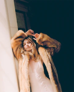 Actor Lily James wearing a fluffy brown faux fur coat and cream dress against a building exterior wall