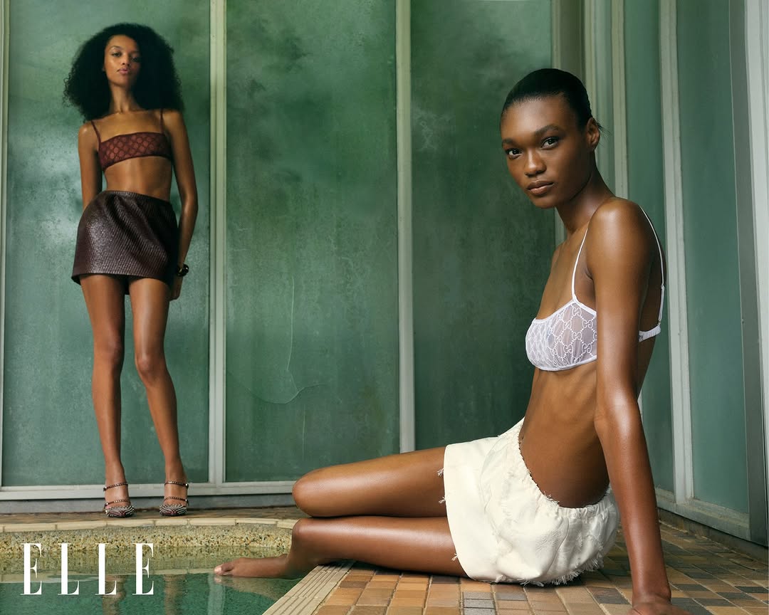 Two Black female fashion models pose indoors; one wears a brown bra top and skirt, the other a white bra top and white skirt.