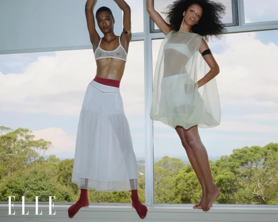 Two slim, dark-skinned women pose barefoot in a window with transparent dresses. A scenic view of trees and sky visible.