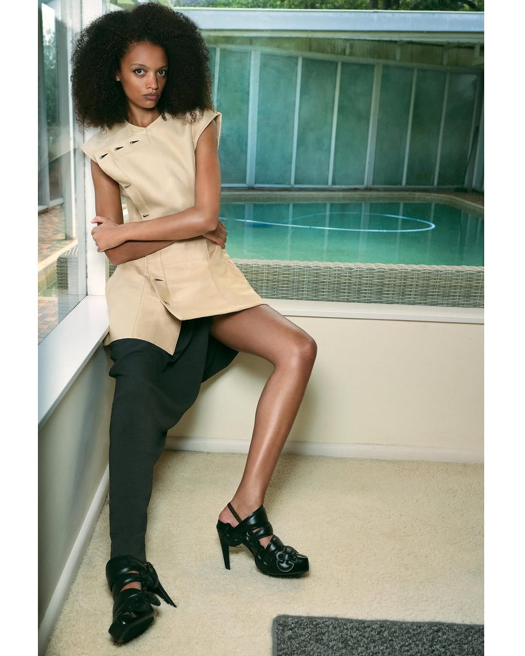 A slender woman with dark, curly hair poses in a beige dress and black high-heeled shoes against an indoor pool backdrop.