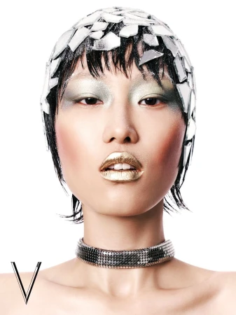 Striking beauty portrait of an Asian woman with shimmery gold lips, dramatic eye makeup, and a broken-mirror cap against a white backdrop.