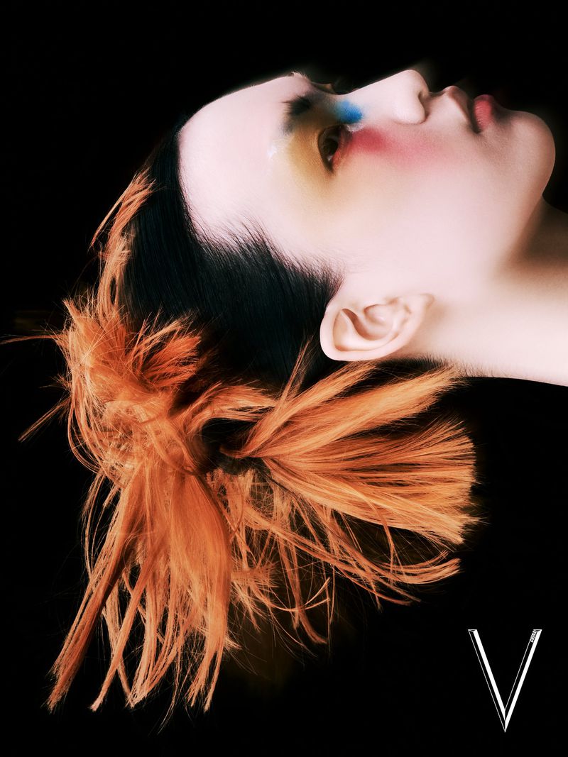 Headshot of a person with dramatic makeup and orange hair against a black background, showing off creative beauty and style.