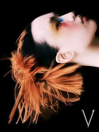 Headshot of a person with dramatic makeup and orange hair against a black background, showing off creative beauty and style.