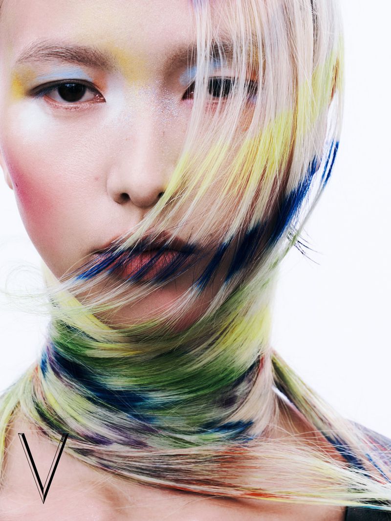 Close-up beauty shot of a person with colorful dyed blonde hair and face makeup against a white background in a studio setting.