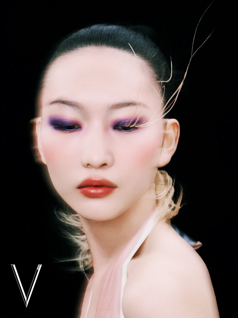 Close-up portrait of a woman with dark hair and dramatic purple eyeshadow, set against a stark black background with the letter V.